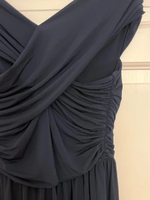 Marinblå långklänning från H&M - Snygg marinblå långklänning från H&M med omlott-detalj upptill och draperad v-ringad överdel. Klänningen är axelbandslös och har ett mjukt, flytande tyg som ger en elegant siluett. Perfekt för dig som vill ha en stilren look med lite extra edge.