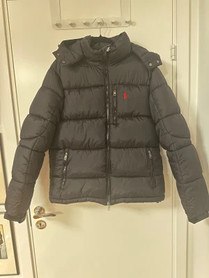 Svart pufferjacka från Polo Ralph Lauren - Inte använt en ända gång eftersom att den var för liten. Svart pufferjacka från Polo Ralph Lauren med röd broderad logga på bröstet. Jackan har hög krage, huva, två sidofickor med dragkedja och en bröstficka med dragkedja. Materialet är syntet och jackan har en quiltad design som ger en schysst volym. Perfekt jacka för kyliga vinterdagar.