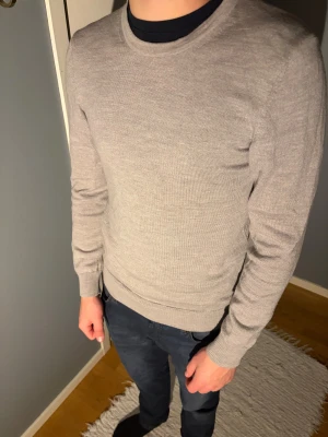 J.lindeberg  - En stilren ljusgrå stickad tröja med rund hals och ribbade muddar. Tröjan har en normal passform och är långärmad, perfekt för lager-på-lager. Materialet känns mjukt och lätt, vilket gör den skön att bära under höst och vinter. 100% marinull