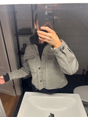 Grå  jeansjacka med broderi - Säljer en grå croppad jeansjacka med snygga broderade detaljer på bröstfickorna och ärmsluten. Utgått ur sortimentet. Som ny. 