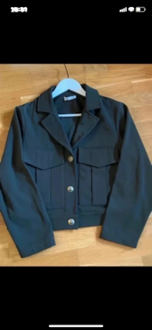 Mörkgrön jacka/ kavaj med fickor - Snygg mörkgrön croppad kavaj/jacka från Design by Si  med klassisk krage och stora bröstfickor. Jackan har långa ärmar och knäpps med tre silvriga knappar framtill. Väldigt snyggt och elegant plagg! Nyskick!! Inga skador eller anmärkningar. Färgen är som på första bilden. 