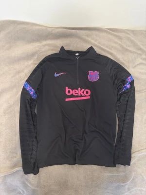FC Barcelona träningströja och byxor - Svart långärmad FC Barcelona träningströja från Nike med halv dragkedja, klubbmärke på bröstet och rosa/blå detaljer. Materialet är lätt och andas, perfekt för fotbollsträning. PRIS KAN DISKUTERAS 
