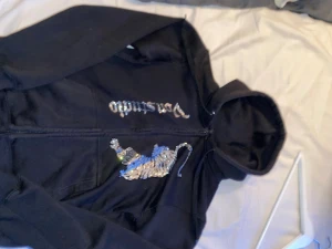 Svart hoodie Von Dutch med strass-tiger - Cool svart hoodie från Von Dutch med dragkedja och huva. På framsidan finns en glittrig tiger i strass och Von Dutch-loggan i silvriga stenar. Perfekt statement-plagg för dig som gillar att sticka ut. Skön passform och snygga detaljer.