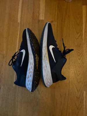Svarta Nike sneakers med vit sula - Säljer ett par svarta Nike sneakers med vit sula och stor vit swoosh på sidan. Ovandelen är i mesh och har snörning. Sulan är tjock och har ett mönstrat grepp undertill, perfekt för löpning eller träning. Snygg och sportig design som funkar till streetwear.