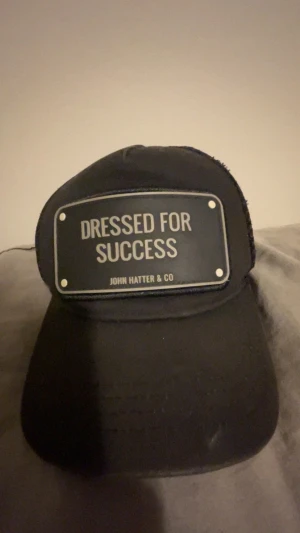 Svart keps John Hatter & Co - Svart keps från John Hatter & Co med en stor metallskylt framtill där det står 'Dressed for Success'. Klassisk böjd skärm och justerbar baksida. Snygg och stilren accessoar som sticker ut.