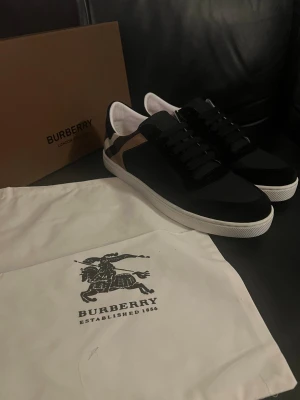 Burberry skor - Snygga svarta sneakers från Burberry med klassisk rutig panel i beige, svart och brunt på sidan. Skorna har vit platt sula, rund tå och svarta snören. Materialmix av skinn och textil ger en lyxig känsla. Perfekt för dig som gillar stilrena och exklusiva sneakers.