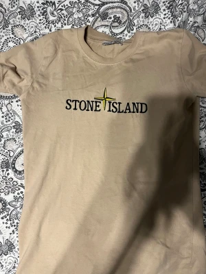 Beige Stone Island t-shirt med broderi - Snygg beige t-shirt från Stone Island med broderad logga och gul kompass på bröstet. Klassisk rund hals och korta ärmar. Tillverkad i mjuk bomull som känns skön mot huden. Perfekt för dig som gillar streetwear och stilrena plagg.   Pris kan diskuteras…