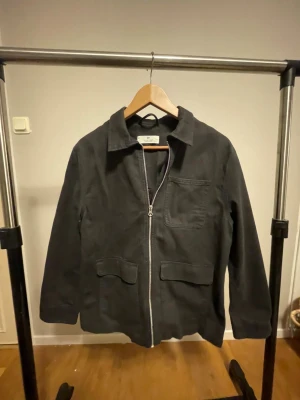 Svart overshirt från H&M med dragkedja - Svart overshirt från H&M i storlek 152. Jackan har en clean look med dragkedja framtill, klassisk krage och tre fickor. Materialet är mjukt och känns som bomull, perfekt för lager-på-lager. Passformen är normal och ärmarna är långa.