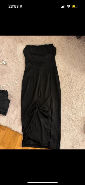 Svart axelbandslös långklänning - Elegant svart axelbandslös långklänning med markerad midja och snygga sömdetaljer på livet. Klänningen har en glittrig kant upptill och en diskret slits framtill. Stängs med dragkedja i ryggen. Perfekt för dig som vill sticka ut med en klassisk och stilren look.