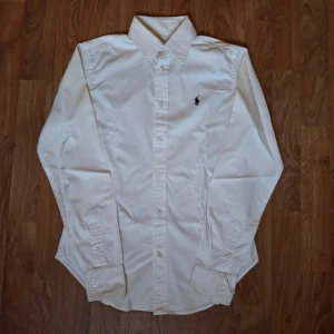 Vit skjorta från Ralph Lauren Sport - Vit skjorta från Ralph Lauren Sport med klassisk krage och knappar framtill. Broderad mörkblå logga på bröstet. Skjortan är långärmad och har manschetter med knappar. Tillverkad i mjuk bomull med lite stretch för extra komfort.