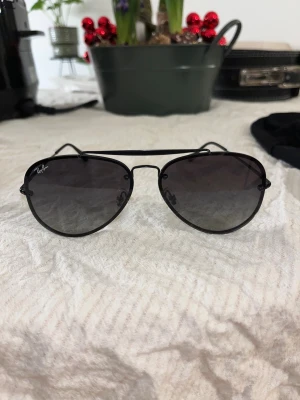 Svarta Ray-Ban aviator solglasögon - Snygga svarta Ray-Ban aviator solglasögon med tunna metallbågar och mörka tonade glas. Klassisk pilotform som ger en cool vibe. Kommer med original Ray-Ban fodral i svart skinnimitation. Perfekta för dig som vill ha en tidlös och ikonisk look.