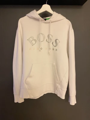 Vit hoodie från Hugo Boss - Vit hoodie från Hugo Boss med stor BOSS-logga på bröstet i silver. Tröjan har huva med snörning, magficka och långa ärmar. Mjuk bomullskvalitet och klassisk passform, perfekt för chill dagar. Snygg och enkel design som funkar till jeans eller joggers. Tröjan har en liten fläck på axeln (se bild)