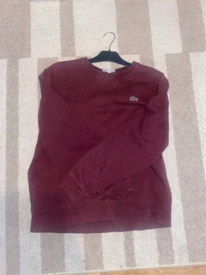 Vinröd och marinblå sweatshirt Lacoste - Två stilrena sweatshirts från Lacoste, en i vinrött och en i marinblått. Båda har klassisk rund hals, långärmad design och den ikoniska krokodilloggan broderad på bröstet. Materialet är mjuk bomull och passformen är normal, perfekt för chill dagar.