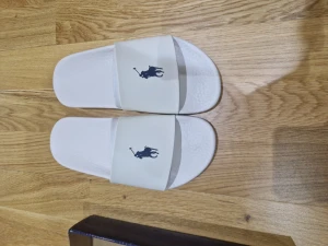 Vita sandaler Polo Ralph Lauren strl 37 - Fräscha vita sandaler från Polo Ralph Lauren i storlek 37. De har en bred rem med den klassiska Polo-loggan i marinblått och en platt sula med mönstrad undersida för extra grepp. Perfekta för sommaren och chill dagar vid poolen. Kom med pris förslag