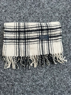 Rutig ullhalsduk från Burberry - Klassisk halsduk från Burberry i mjuk lammull. Vit botten med svart rutigt mönster och tvinnade fransar i ändarna. Perfekt accessoar för att lyfta din outfit. 