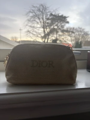 Beige necessär från Dior med stjärndetalj - Snygg beige necessär från Dior med glittrig finish och guldig dragkedja. Väskan har en stjärnformad Dior-tag på sidan och är perfekt för smink eller småprylar. Stilren och lyxig design som sticker ut i väskan eller på hyllan.