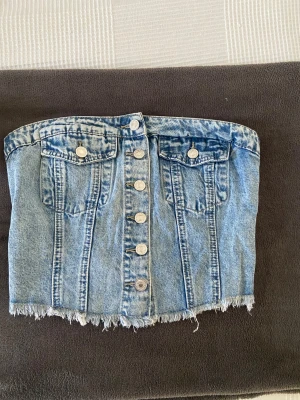 Jeans korsett/topp - Säljer en snygg tubtopp i ljusblå denim med silverfärgade knappar framtill och två bröstfickor. Toppen har fransad nederkant och smockad rygg för skön passform. Perfekt för dig som gillar denim.