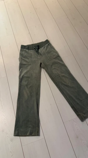 Gröna velourbyxor Juicy Couture S - Snygga gröna velourbyxor från Juicy Couture i storlek S. Byxorna har bred resårmidja med snörning, stora fickor fram och bak med metallknappar, samt broderad logga på ena fickan. Modellen är rak och loose fit för chill vibe. Nypris 1200kr