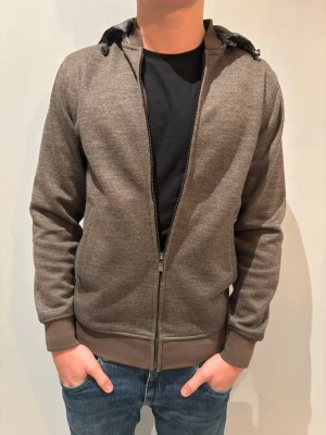 Brun Massimo dutti - Snygg brun hoodiejacka med grå huva och dragkedja framtill. Jackan har ribbade muddar vid ärmslut och nederkant, samt praktiska fickor på sidorna. Perfekt för chill dagar och enkel att matcha med jeans eller joggers. Modellen är 175 cm