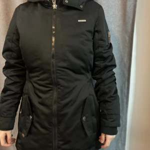 Svart parkajacka från Marikoo XS - Svart parkajacka från Marikoo med huva, dragkedja och knappar framtill. Jackan har långa ärmar, två stora fickor med lock och knapp, samt en snygg detalj på ärmen. Insidan är fodrad med mjuk fleece för extra värme. Perfekt för kalla dagar. Andvänd fåtal gånger säljer för 600kr
