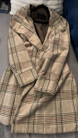 Beige rutig kappa från Giovanna - Snygg beige kappa från Giovanna i klassiskt rutigt mönster med svarta och röda linjer. Jackan har långa ärmar, knappar framtill. Perfekt för dig som gillar stilrena och tidlösa plagg med lite edge.