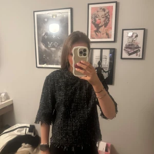 Svartvit rutig blus med fransar - Säljer en svartvit rutig blus från Zara med hög krage och trekvartsärm. Blusen har fransade kanter vid ärmslut och nederkant, samt ett grovt vävt material som ger en cool struktur. Perfekt för dig som gillar statement-plagg med edge.