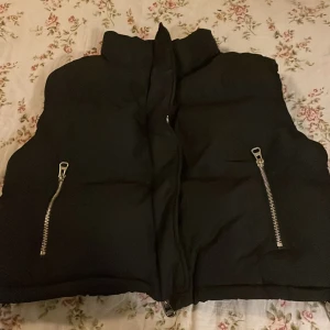 Svart dunväst från FB Sister XS - Svart dunväst från FB Sister i storlek XS. Västen har två stora silvriga dragkedjor framtill och hög krage som ger en cool look. Materialet är mjukt och syntetiskt, perfekt för lager-på-lager under höst och vinter. Passformen är normal och västen är quiltad för extra värme.