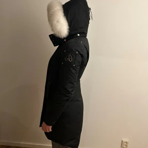 Svart Moose Knuckles jacka med äkta päls - Snygg svart lång vinterjacka från Moose Knuckles med stor vit päls runt huvan. Jackan har dragkedja framtill, flera fickor och logga på ärmen. Perfekt för vintern då den håller värmen extremt bra och jackan är också i väldigt bra skick skulle säga nästintill oanvänd då jag inte använt jackan mer än några få gånger då den blivit för liten för mig (inte jag på bilden) skriv gärna vid frågor och intresse! Säljer för väldigt bra pris då nypris ligger på väldigt mycket mer och jackan är i toppen skic