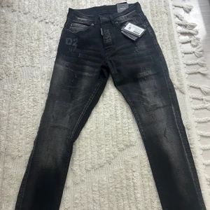 Svarta Skater Jeans från Dsquared2 - Säljer ett par svarta Dsquared2 Skater Jeans med coola slitningar och tryckta detaljer som D2 och lönnlöv. Jeansen har blå insida i fickorna, klassisk femficksdesign och dragkedja. Passformen är slim och materialet är jeans med snygg tvättad look.