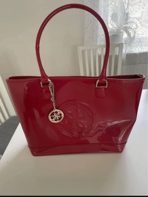 Röd handväska från Guess - Snygg röd handväska från Guess i glansigt skinnmaterial med präglad logga framtill och metallhänge. Väskan har två rejäla handtag och ett rymligt innerfack med dragkedja och mönstrat tygfoder med Guess-loggor. Perfekt för dig som gillar att sticka ut.