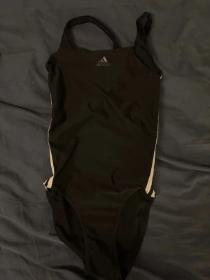  Adidas baddräkt  - Snygg svart baddräkt från Adidas med klassiska vita ränder längs sidorna och Adidas-logga framtill. Helt ny, tog bara bort prislapp då jag var säker på att den skulle användas men den gjorde aldrig det. Storlek 32/xs väldigt fin baddräkt och köpt för 799kr för två år sedan  
