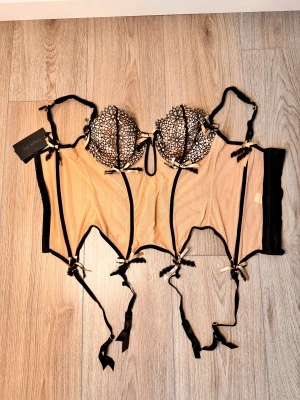 Svart och beige korsett från Patrizia Pepe - Säljer en elegant korsett i svart och beige mesh med blommig spets på kuporna. Den har justerbara axelband, dekorativa rosetter och hakknäppning bak. Perfekt för dig som vill ha en snygg och unik topp med coola detaljer.