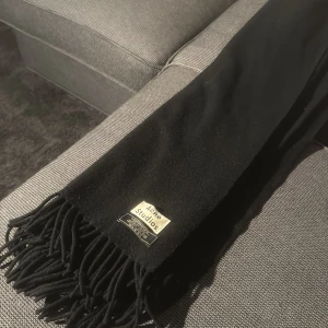 Svart Acne Studios halsduk i ull - Snygg svart halsduk från Acne Studios i mjuk lammull. Klassisk rektangulär form med stilrena fransar i ändarna och en diskret logotyp på ena sidan. Perfekt för att ge din outfit en clean och modern vibe.