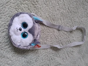 Ty Gear Slush plysch axelväska - Supergullig rund axelväska från Ty Gear med plyschmaterial och motiv av hunden Slush. Väskan är grå och vit med stora blå glittriga ögon och små blå öron som föreställer en varg. Justerbar grå axelrem och mjuk finish, perfekt för att bära småsaker. Köpt utomlands, finns inte i Sverige.