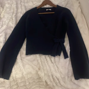 Svart omlott kofta med rosett - Mysig svart omlott kofta med lång ärm och en snygg rosett på sidan. Koftan är stickad och har en lite croppad passform som ger en cool vibe. Perfekt att slänga på sig när det är lite kyligt ute.