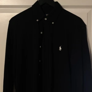 Svart skjorta från Polo Ralph Lauren - Svart långärmad skjorta från Polo Ralph Lauren med klassisk button-down krage och vit broderad logga på bröstet. Skjortan har knappar framtill och är i en stilren, tidlös design som passar till många olika looks.