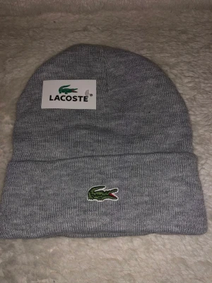 Lacoste Mössa  - Snygg grå mössa från Lacoste med klassisk krokodillogga framtill. Stickad i mjukt material med uppvikt kant för extra stilpoäng. Perfekt för dig som vill ha en clean look och samtidigt visa upp ett ikoniskt märke.