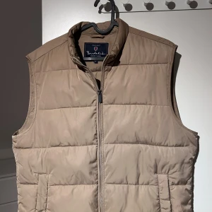 Beige dunväst från Bondelid - Snygg beige dunväst från Bondelid med quiltad design och hög krage. Västen har två sidofickor och dragkedja framtill. Perfekt att slänga över en hoodie för extra värme. Modern och stilren look som funkar till många outfits. Hör av dig vid minsta lilla fråga:)