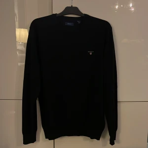 Svart tröja från GANT i XL - Klassisk svart tröja från GANT i storlek XL. Tröjan har rund halsringning, långa ärmar och en liten broderad GANT-logga på bröstet. Perfekt för en stilren och enkel look.