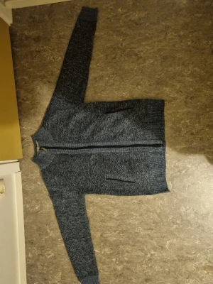 Blå stickad kofta med dragkedja - Mysig blåmelerad stickad kofta med hel dragkedja framtill och två fickor. Insidan har ett rutigt foder i beige, brun och svart som ger extra värme. Ribbstickade muddar vid ärmslut och nederkant. Perfekt för kyliga dagar.