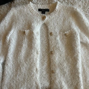 Vit fluffig kofta från Amisu med guldknappar - Supermysig vit kofta från Amisu i storlek S. Koftan är fluffig och stickad med snygga, strukturerade guldknappar framtill. Rund halsringning och långärmad modell, perfekt för lager-på-lager och att ge din outfit en lyxig touch.
