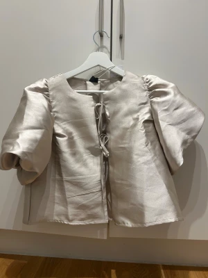 Champagnefärgad satin blus med puffärm - Säljer en elegant blus i champagnefärgad satin med puffiga korta ärmar och snygg snörning framtill. Blusen har en rund halsringning och glansigt material som ger en lyxig känsla. Perfekt för dig som vill sticka ut med stilrena detaljer.