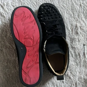 Christian Louboutin svarta sneakers med nitar - Svarta sneakers från Christian Louboutin med ikoniska röda sulor och svarta nitar över tån och hälen. Skorna har snörning, rund tå och är tillverkade i mocka och skinn. Insidan är beige och har märkets logga tryckt i rött.