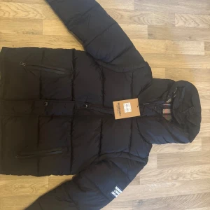 Svart pufferjacka från Burberry - Ny Burberry dunjacka – Storlek S (kan göras om till väst)  Säljer en helt ny och oanvänd Burberry dunjacka i storlek S. Jackan är i perfekt skick, med tag kvar, och är både varm och stilren. Ärmarna är avtagbara, så den kan enkelt göras om till en väst – perfekt för både kallare och mildare dagar. 