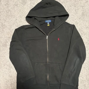 Svart hoodie från Polo Ralph Lauren XL - Svart hoodie från Polo Ralph Lauren i storlek XL med dragkedja och huva. Klassisk röd broderad logga på bröstet och praktisk känguruficka framtill. Mjuk bomullskänsla och stilren look, perfekt för chill dagar.