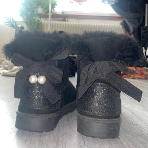Svarta fluffiga boots med rosett och pärlor - Säljer ett par svarta boots med glittrig häl och stora rosetter bak med pärldetaljer. Skorna är gjorda i mockamaterial och har ett väldigt mjuk foder. Saknas tyvärr pärlor på ena skon. Storlek: 37/38 (står 37 men skulle säga att de är lite större)
