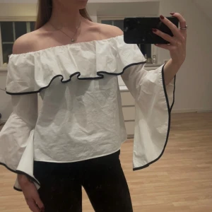 Vit offshoulder blus med volang - Säljer en jätte snygg offshoulder blus med volang runt axlarna och svarta kantdetaljer. Blusen har vida, långa ärmar och är tillverkad i ett lätt bomullstyg. Passar xs-m då den är väldigt stretchig. Nyskick❤️