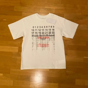 Maison Margiela vit t-shirt med print - Vit t-shirt från Maison Margiela med ikoniskt siffror-print och text i svart och rött på framsidan. Klassisk rund hals och korta ärmar. Tillverkad i mjuk bomull, med diskreta stygn på ryggen som signaturdetalj. Snygg och clean design med edgy vibe.