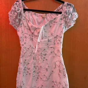 Vit blommig kortklänning Love triangle - Superfin vit kortklänning från Love triangle med broderade blommor och puffiga korta ärmar i mesh. Klänningen har söt snörning framtill och romantiska detaljer. Perfekt för dig som gillar en feminin och trendig stil.