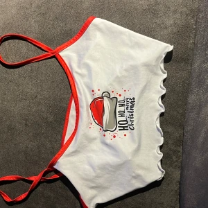 Vit croppad linnetopp med jultryck - Säljer en vit croppad linnetopp med röda kantband och smala axelband. Framsidan har ett tryck med tomteluva och texten 'Ho Ho Ho Merry Christmas'. Toppen har vågig volangkant nedtill och är perfekt för julens alla tillfällen.